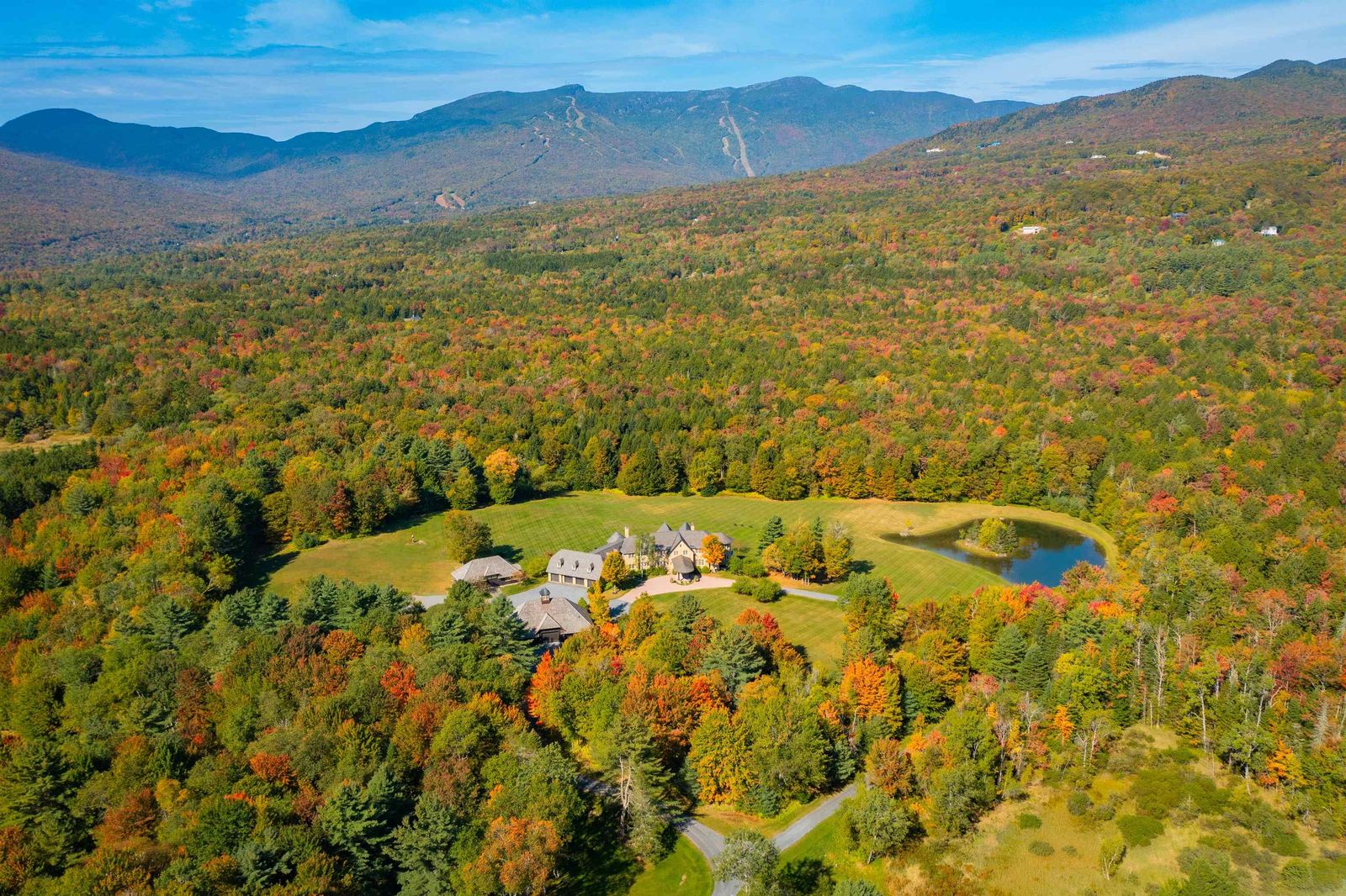 Aerial view of Pristine Meadow at 2575 Weeks Hill Road in Stowe, Vermont, showing the stone residence, outbuildings, and swimming pond in a meadow surrounded by fall woodland, with ski trails on the mountain behind